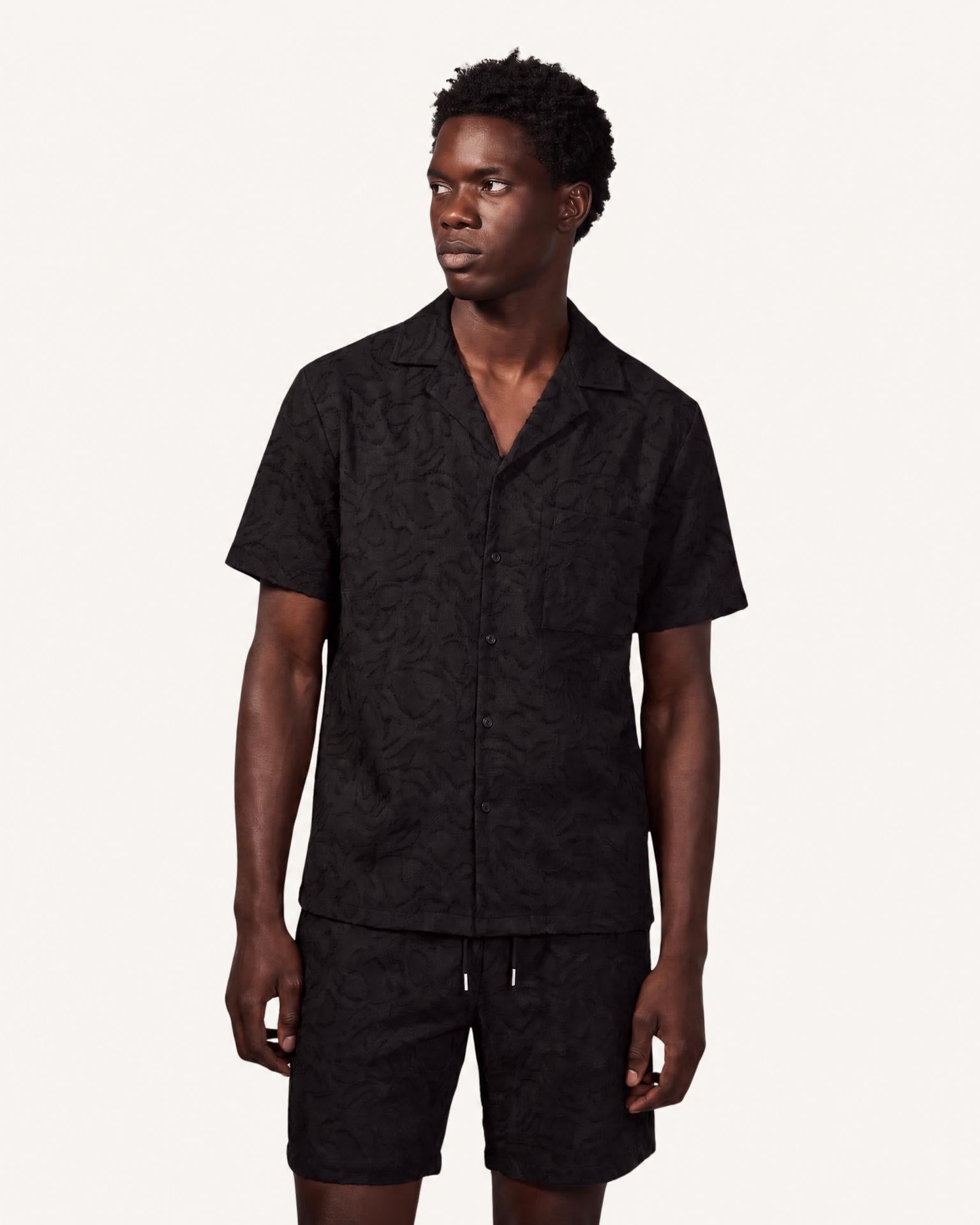 A man modeling a black short-sleeve button-up shirt and matching shorts set with a subtle tonal damask pattern, photographed against a solid off-white background.