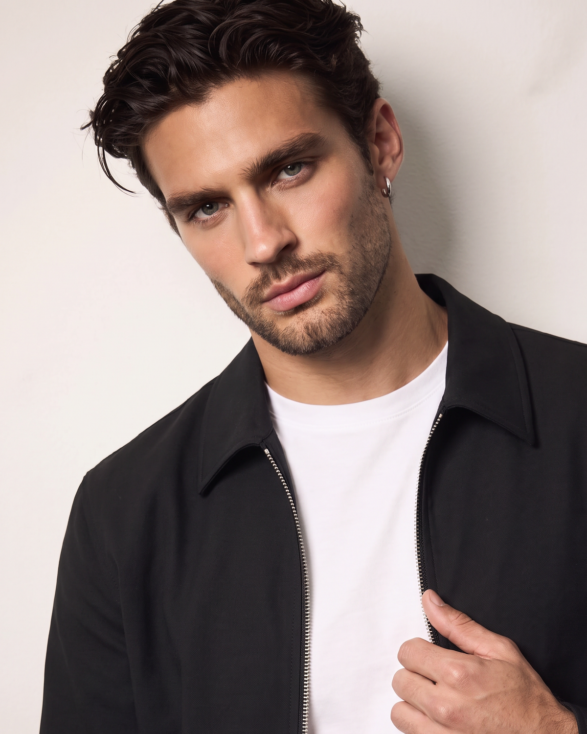 A man wearing a black zip-up jacket over a white t-shirt, posing against a light off-white background.