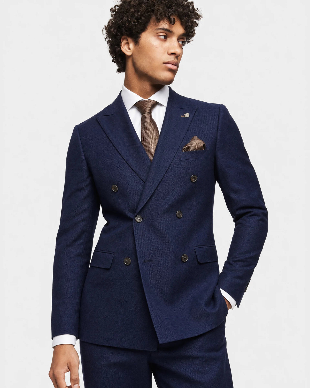 A man wearing a navy blue double-breasted suit with a white dress shirt, brown tie, and matching pocket square, photographed against a light gray studio background.
