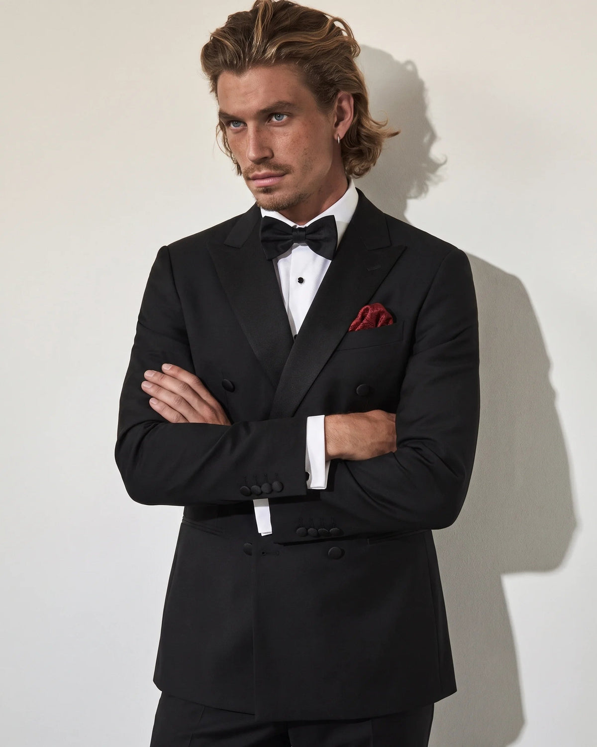 A man wearing a black double-breasted tuxedo jacket with a black bow tie, white dress shirt, and a red pocket square, standing with arms crossed against a light gray wall.