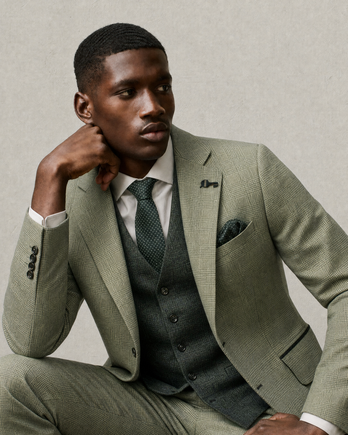 A man wearing a light sage green three-piece suit with a matching vest and tie, posed against a neutral textured gray background.